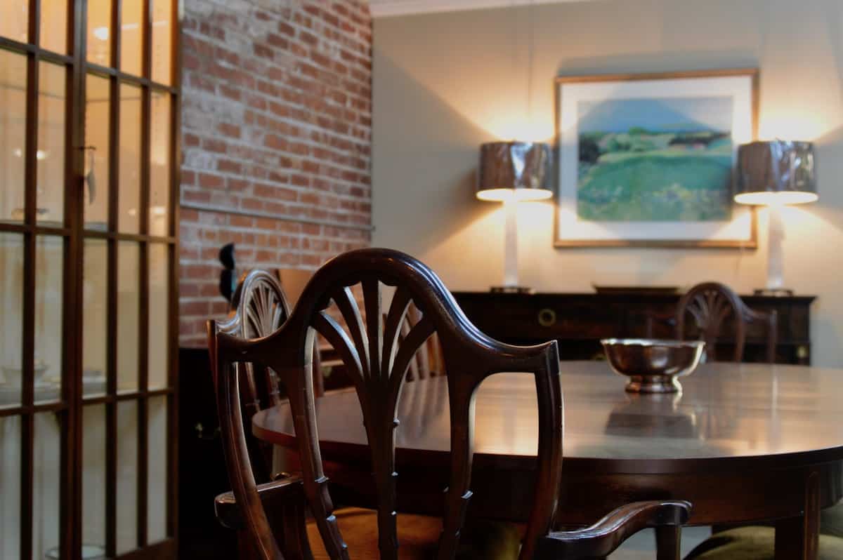 Warm dining room interior with a round wood table, carved chairs, exposed brick wall, and soft lamp lighting