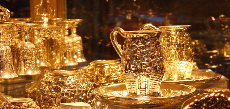 Close-up of ornate gold-toned metal pitchers, cups and decorative vessels with intricate engraved designs, warm golden lighting against a dark background
